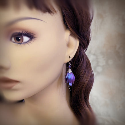 Flower-shaped earrings made with iridescent blue and purple tulip drops. The earrings are adorned with antiqued silver-colored leafy bead caps and antiqued silver-colored pins dangling beneath the flowers resembling stamen. Each flower sits a matching iridescent Czech glass bead on top. Shown on a mannequin for scale