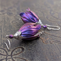 Flower-shaped earrings made with iridescent blue and purple tulip drops. The earrings are adorned with antiqued silver-colored leafy bead caps and antiqued silver-colored pins dangling beneath the flowers resembling stamen. Each flower sits a matching iridescent Czech glass bead on top.