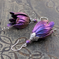 Flower-shaped earrings made with iridescent blue and purple tulip drops. The earrings are adorned with antiqued silver-colored leafy bead caps and antiqued silver-colored pins dangling beneath the flowers resembling stamen. Each flower sits a matching iridescent Czech glass bead on top.