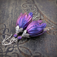 Flower-shaped earrings made with iridescent blue and purple tulip drops. The earrings are adorned with antiqued silver-colored leafy bead caps and antiqued silver-colored pins dangling beneath the flowers resembling stamen. Each flower sits a matching iridescent Czech glass bead on top.
