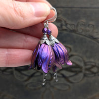Flower-shaped earrings made with iridescent blue and purple tulip drops. The earrings are adorned with antiqued silver-colored leafy bead caps and antiqued silver-colored pins dangling beneath the flowers resembling stamen. Each flower sits a matching iridescent Czech glass bead on top.