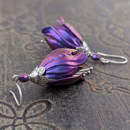 Flower-shaped earrings made with iridescent blue and purple tulip drops. The earrings are adorned with antiqued silver-colored leafy bead caps and antiqued silver-colored pins dangling beneath the flowers resembling stamen. Each flower sits a matching iridescent Czech glass bead on top.