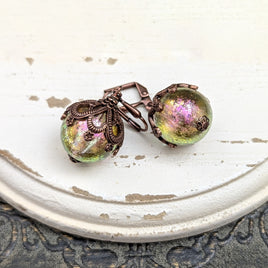 Handmade earrings with large hand-colored translucent beads with an iridescent color-shifting finish in bright pink and green. The earrings are capped with intricately detailed floral filigree in an antiqued copper finish.