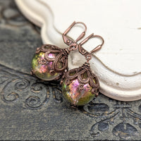 Handmade earrings with large hand-colored translucent beads with an iridescent color-shifting finish in bright pink and green. The earrings are capped with intricately detailed floral filigree in an antiqued copper finish.