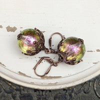 Handmade earrings with large hand-colored translucent beads with an iridescent color-shifting finish in bright pink and green. The earrings are capped with intricately detailed floral filigree in an antiqued copper finish.