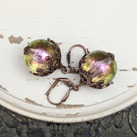 Handmade earrings with large hand-colored translucent beads with an iridescent color-shifting finish in bright pink and green. The earrings are capped with intricately detailed floral filigree in an antiqued copper finish.
