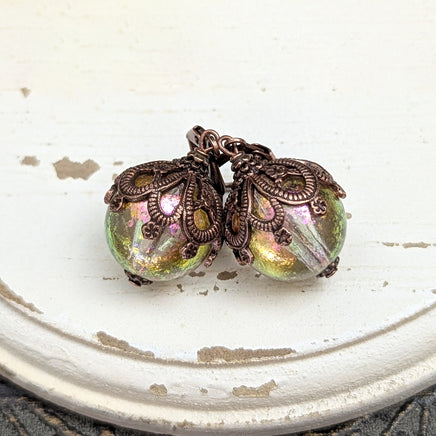 Handmade earrings with large hand-colored translucent beads with an iridescent color-shifting finish in bright pink and green. The earrings are capped with intricately detailed floral filigree in an antiqued copper finish.