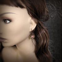 Handmade earrings with large hand-colored translucent beads with an iridescent color-shifting finish in bright pink and green. The earrings are capped with intricately detailed floral filigree in an antiqued copper finish. Photographed on a mannequin for scale.