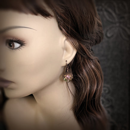 Handmade earrings with large hand-colored translucent beads with an iridescent color-shifting finish in bright pink and green. The earrings are capped with intricately detailed floral filigree in an antiqued copper finish. Photographed on a mannequin for scale.
