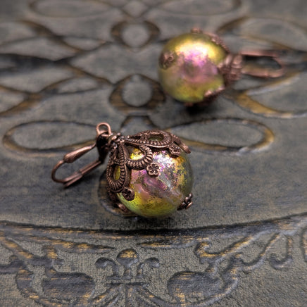 Handmade earrings with large hand-colored translucent beads with an iridescent color-shifting finish in bright pink and green. The earrings are capped with intricately detailed floral filigree in an antiqued copper finish.